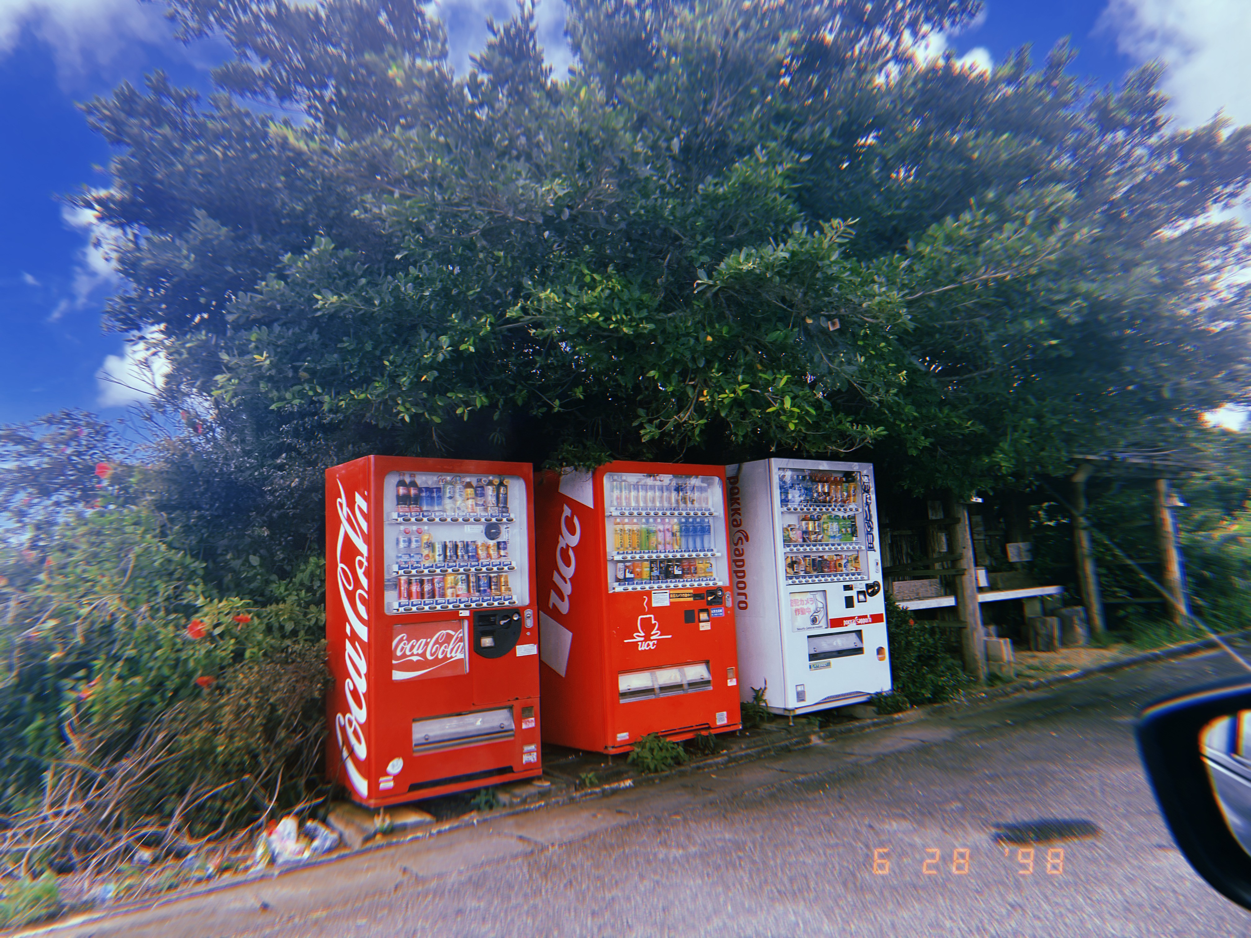Impressionist-style painting of a landscape with a bright blue sky full of clouds, a green field with yellow flowers, a large tree in the foreground, and a castle and small trees in the distance in front of a small mountain